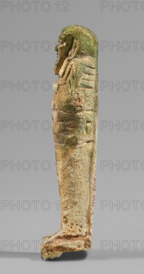 Ushabti, Wigs, funerary figurines, hoes, inscriptions, grave goods. Mummy-shaped figurine, standing on a base, supported at the back by a flat, wide pillar. Arms crossed on the chest, only the hands visible from under the shroud; in both hands are hoes, and in the right hand a rope on which a basket is slung over the left shoulder. Engraved tools. On the head is a smooth, three-part wig merging at the back into a pillar separated from it by a thin horizontal line; the rear strand is significantly longer than the front strands. Ears uncovered. Large bulging eyes, wide nose and mouth. Long straight beard. Engraved inscription on the front of the figurine's legs. T-shaped. 664–332 BC; Late Period, Egypt. Shabti. Height 10.8 cm, width 3 cm., A slender upright statuette viewed in profile, showing a standing human figure with a stylized wig or headdress, a beard, and elongated body proportions; the surface shows a weathered, pitted texture and areas of encrustation. The visible colors includ...