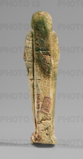 Ushabti, Mummy-shaped funerary figurine, standing on a base and supported at the back by a flat, wide pillar. Arms crossed on the chest with only the hands visible from under the shroud; each hand holds hoes and the right hand also holds a rope from which a basket is slung over the left shoulder. Engraved tools. Smooth three-part wig on the head merging at the back into the pillar separated by a thin horizontal line; rear strand significantly longer than the front strands. Ears uncovered; large bulging eyes, wide nose and mouth; long straight beard. Engraved inscription on the front of the figurine's legs. T-shaped. Date: 664–332 BC, Late Period, Egypt. Shabti. Height 10.8 cm, width 3 cm., A weathered vertical statuette viewed from the back, with a rounded head and elongated body tapering toward the base, showing a textured surface with worn areas and small pits; the object displays a mix of green and beige tones with patches of darker green and lighter sandy beige, plus accents of bro...