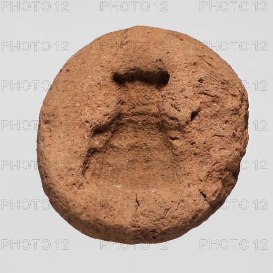 A mold for making an amulet — Bes mask, Bes myth.; mask amulet casting; mold; height 0.4 cm; 664–332 BC Late Period, Egypt., A roughly circular, textured object with an uneven, cratered indentation in its center, showing a coarse, grainy surface with small pits and irregularities; the object appears in shades of brown with specks of lighter and darker brown and tiny flecks of off-white, set against a pale gray background.