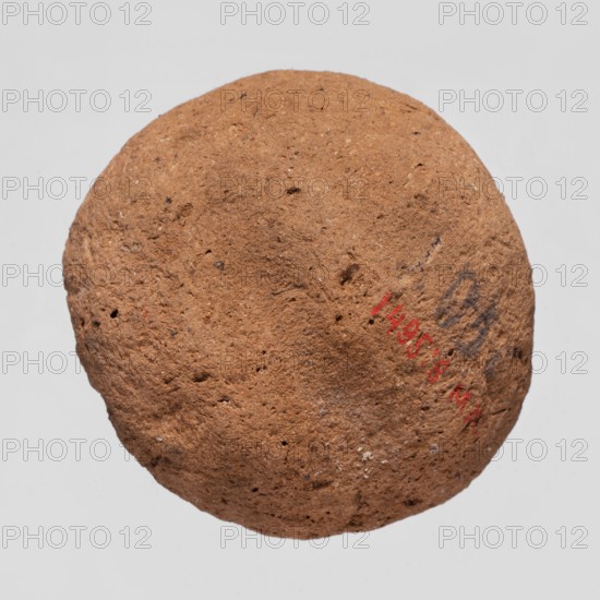 A mold for making an amulet - Bes mask, Bes myth., mask amulet casting; mold, height 0.4 cm; 664–332 BC, Late Period, Egypt., A roughly spherical, textured object with a coarse, porous surface in shades of brown and tan, showing small pits and irregularities; a few small darker brown and black specks are scattered across the surface, and there is a faint area of darker grayish markings on one side against a plain light gray background.