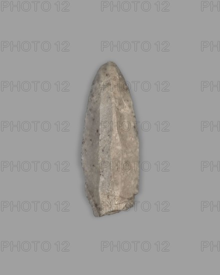Javelin head, militaria, arrowheads, ranged weapons, material culture, Eastern Gaulish scenography, 4th millennium BC, Southern Mesopotamia, Sumer, weapon, weight 10 g, A roughly elongated, pointed stone object with faceted surfaces sits against a uniform gray background; the stone shows a mix of light gray, beige, and pale brown tones with small darker speckles, uneven edges, and visible flake scars along its sides, and its surfaces vary between smoother, slightly reflective planes and rougher, more textured areas.