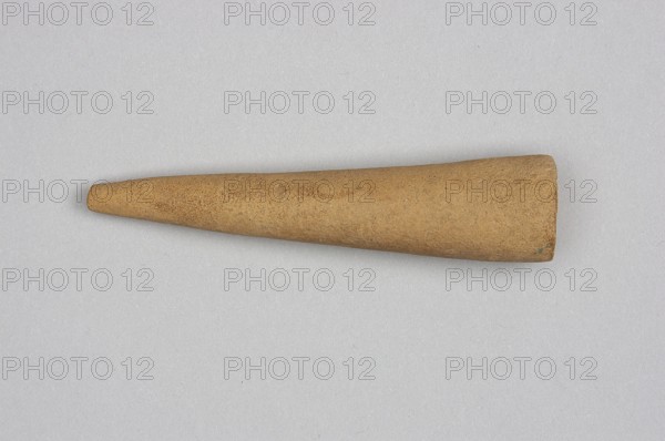 Mosaic cone; decorative architectural element from facades of cult buildings. Mosaic cones; Sumer, Uruk culture, 4th millennium BC. Southern Mesopotamia; element of architectural decoration., A single tapered cylindrical object lies horizontally on a light gray background, appearing textured and sandy with shades of tan and brown; the object narrows to a rounded tip on one end and broadens to a flattened, slightly uneven end on the other, with subtle darker speckles and small irregular surface marks visible across its surface.
