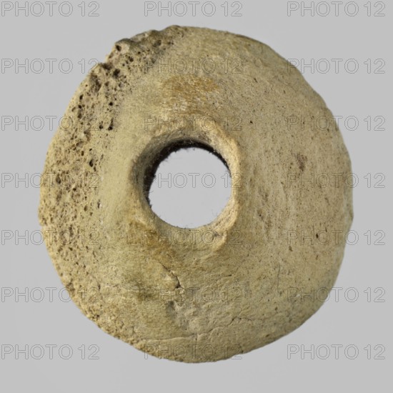 Spindle whorl, weaving tool, material culture, tools, Eastern Gaulish scene, 4th millennium BC, Sumer, Southern Mesopotamia, tool, weight 6.08 g, A roughly circular, doughnut-shaped object with a central hole, showing a textured, pitted surface on one side and a smoother surface on the other, with visible cracks and irregularities. The object appears in shades of beige, tan, and light brown against a light gray background.