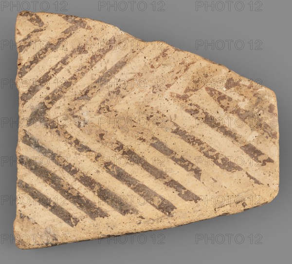 Fragment of a bowl spout, unknown northern Mesopotamian workshop; painted pottery with geometric motifs; Near Eastern pottery; c. 6900–c. 6500 BC; Hassuna culture, Northern Mesopotamia; vessel; weight 28 g, A triangular fragment of pottery with a beige base color and a pattern of diagonal dark brown stripes across its surface, showing irregular edges and some chipping, small scattered darker speckles and patches, and an overall worn, slightly rough texture against a neutral gray background.