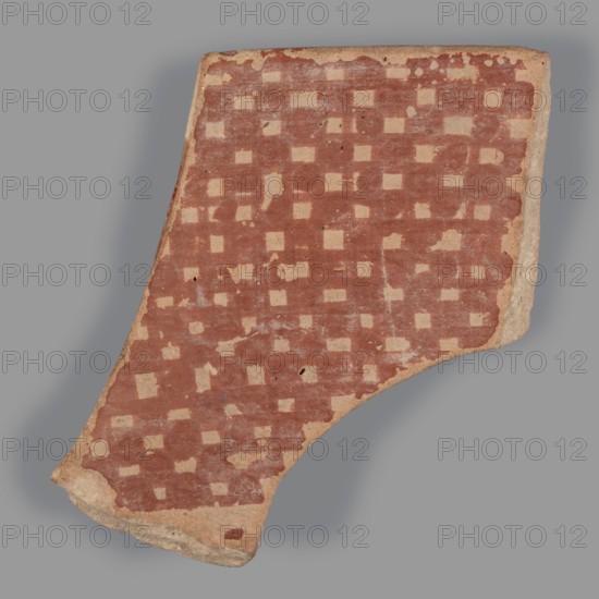 Fragment of a bowl, unknown Northern Mesopotamian workshop. Checkerboards, geometric motifs, painted pottery; Halaf culture c. 5200–c. 4500 BC; earlier dating c. 6500–c. 5500 BC. Northern Mesopotamia Iraq/Syria/Turkey. Pottery fragment: wall of a bowl with preserved thin rim. Before firing, decoration of geometric motifs was painted with ferrous clay on both sides over a layer of dried, heavily smoothed slip/glaze; after firing the painted motifs are red, background cream on the outside and light brown on the inside. Outer decoration: banded composition from top downward — a wide horizontal band, then alternating rows of single dots and two continuous thin lines, below which a single thin line with superimposed irregular ovals. Inner surface covered with a fine red checkered motif. Material: pottery. Dimensions: height 4.7 cm, width 4.6 cm, depth 0.5 cm; weight 10 g., A broken shard of pottery photographed against a gray background, showing a worn surface patterned with a grid of light...