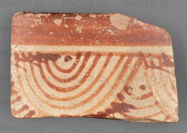 Fragment of a bowl, unknown Northern Mesopotamian workshop, painted pottery, geometric motifs, bowls, fish scale ornament. Fragment of the wall of a bowl or cup with a preserved thin, pointed rim. Decoration painted on both sides, composed of geometric motifs. On the outside, the composition consists of from the top: a wide horizontal band separated by a narrow ribbon of light background from another band filled with groups of seven diminishing semicircular lines and dots arranged like fish scales. On the inside, the decoration was limited to a narrow strip running under the rim. c. 6500–c. 5500 BC; Halaf culture c. 5200–c. 4500 BC earlier dating, Northern Mesopotamia Iraq, Syria, Turkey. Pottery fragment; height 3 cm, width 4.6 cm, depth 0.4 cm, weight 8 g., A broken flat piece of pottery with a background of beige and a pattern painted in reddish-brown; the top edge shows a solid reddish-brown band with areas of worn beige patches, below which a thin beige stripe separates the band f...