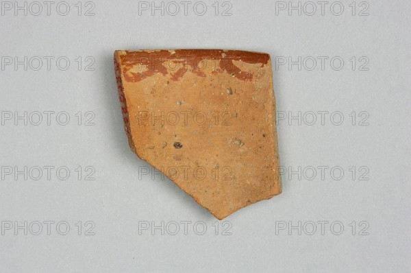 Fragment of a bowl spout, 6th millennium BC, Northern Mesopotamia, vessel, A fragment of pottery with an irregular, angular shape sits on a light gray background; the fragment's surface is mainly a warm orange-brown with speckled inclusions and a slightly rough texture, the top edge shows a band of darker reddish-brown with a few lighter patches, and one side has a narrow strip showing a red hue.
