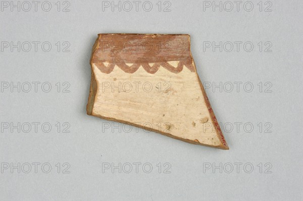 Fragment of a bowl, unknown Northern Mesopotamian workshop; painted pottery with geometric motifs, bowls, Near Eastern pottery. Fragment of the wall of a bowl or cup with a preserved pointed rim. On the outside, the decorative composition consists of from the top: a wide band of diagonally intersecting parallel lines, terminated at the bottom by a thick line. Below is a fragment of another band filled with a row of triangles or rhombuses filled with diagonally intersecting lines. On the inside, the decoration is limited only to the upper part of the vessel and consists of a wide band terminating at the bottom with a wavy line. c. 6500–c. 5500 BC; Halaf culture ca. 5200–ca. 4500 BC, Northern Mesopotamia Iraq/Syria/Turkey. Pottery fragment; height 3.8 cm, width 3.8 cm, depth 0.48 cm, weight 8 g., A broken shard of pottery with an irregular triangular shape rests on a light gray background; the shard shows a smooth off-white surface with small speckles and a darker reddish-brown rim along...