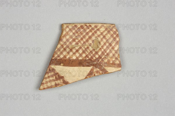 Fragment of a bowl, unknown Northern Mesopotamian workshop, painted pottery with geometric motifs, bowls, Eastern Gaulish Scenery. Fragment of the wall of a bowl or cup with a preserved pointed rim. On the outside, the decorative composition consists of from the top: a wide band of diagonally intersecting parallel lines, terminated at the bottom by a thick line. Below is a fragment of another band filled with a row of triangles or rhombuses filled with diagonally intersecting lines. On the inside, the decoration is limited only to the upper part of the vessel and consists of a wide band terminating at the bottom with a wavy line. c. 6500–c. 5500 BC; Halaf culture ca. 5200–ca. 4500 BC, Northern Mesopotamia Iraq/Syria/Turkey. Pottery fragment; height 3.8 cm, width 3.8 cm, depth 0.48 cm, weight 8 g., A broken shard with an angled triangular shape lies on a light gray background; the shard shows a checkered pattern in reddish-brown and beige across its upper area, a horizontal reddish-brow...