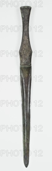 Short sword, unknown Luristan workshop. A short sword with a cast blade and a mushroom-shaped hilt with a rimmed mount; the raised edges served to hold the lost hilt casing, which may have been made of bone, wood, horn, or stone. In the lower part of the casing, two low, crescent-shaped sheet-metal protrusions are bent inward to secure the casing. The hilt flows directly into a double-edged blade with a centric point. Behind the short, rectangular, double-blunt base, the blade tapers evenly toward the tip. A slightly convex ridge runs through the center of the blade, which splits toward the base in its upper part. Luristan; Iran; 9th–6th century BC, Iron Period II late 2nd millennium–first half of 1st millennium BC. Weapon. Height 40.6 cm; width 3.4 cm; depth 2 cm; weight 208 g., A single elongated object resembling a narrow blade and handle is shown vertically against a plain light background, with the handle wider at the top and tapering toward the middle, transitioning to a straight...