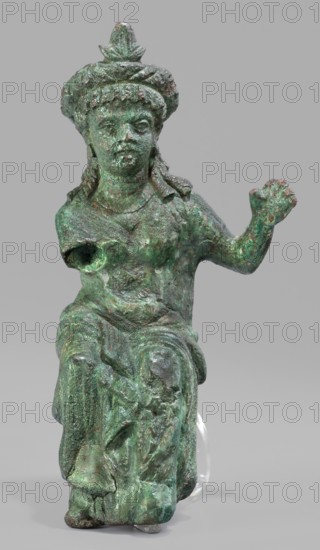 Figurine of the goddess Aphrodite-Isis; maker, Egyptian deities; wigs; crowns; female figurines; Aphrodite; Isis; Egyptian art; 1st–2nd century Roman period; origin: Egypt possibly Rome?; figurine; height 11.1 cm, width 5.3 cm, depth 5 cm, A seated metal figurine with a weathered green patina, showing a seated figure wearing a rounded headdress with a pointed crest and textured band, with long hair or braided elements falling to the shoulders; the face has worn features and a neutral expression, and one arm is raised with an open hand while the other arm is broken off at the shoulder; the figure wears draped garments with visible folds and textured surface, and the lower body and legs are covered by the robe with signs of corrosion and rough surface, set against a plain gray background with small areas of darker and brownish discoloration on the metal.