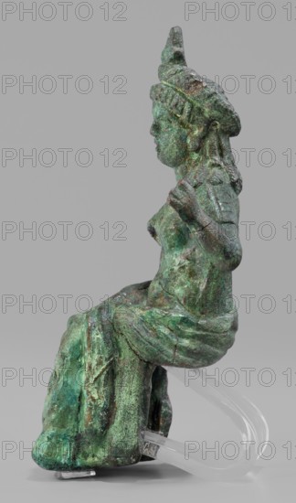 Figurine of the goddess Aphrodite-Isis, Egyptian deities, wigs, crowns, female figurines, Aphrodite mythology, Isis mythology, Egyptian art, 1st–2nd century Roman period, Egypt ? Rome?, figurine, height 11.1 cm, width 5.3 cm, depth 5 cm, A green and brown weathered metal figurine of a seated person shown in profile against a gray background, with a textured surface and areas of darker and lighter green patina, brown corrosion, defined facial features, a headdress with a pointed top, long hair or braids at the back, an arm bent at the elbow with a closed hand near the chest, draped clothing over the legs with visible folds, and a clear curved support under the figure.