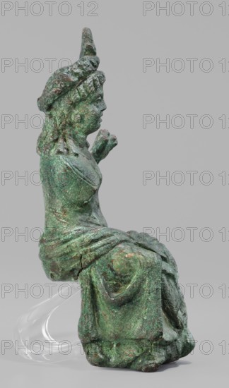 Figurine of the goddess Aphrodite-Isis, Egyptian deities, wigs, crowns, female figurines, Aphrodite, Isis, Egyptian art, 1st–2nd century Roman period, Egypt possibly Rome, terracotta figurine, height 11.1 cm, width 5.3 cm, depth 5 cm, A greenish metal statuette of a seated figure shown in profile against a plain light gray background, with a textured patina showing variations of green, darker green, and traces of brown and copper tones; the figure wears a headdress with a twisted or braided element and has long hair draped down the back; the torso shows a draped garment with visible folds and layered fabric over the lap and legs; one arm is bent with the hand raised near the chest while the other arm is less distinct; the statuette rests on a clear curved support beneath the seat that is translucent and glossy.