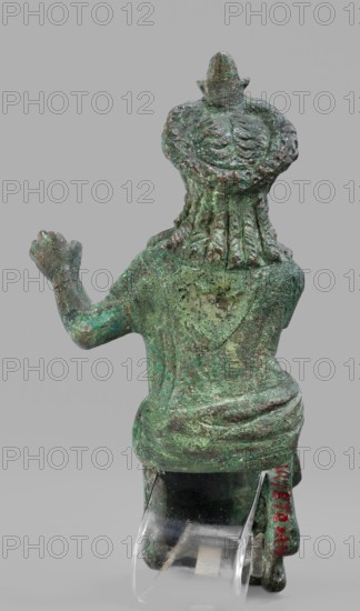 Figurine of the goddess Aphrodite-Isis, Egyptian deities, wigs, crowns, female figurines, Aphrodite mythology, Isis mythology, Egyptian art; 1st–2nd century, Roman period; Egypt possibly Rome?; terracotta figurine, height 11.1 cm, width 5.3 cm, depth 5 cm, A weathered green and brown metal figurine viewed from the back, showing textured hair or headdress details and a rounded upper back with draped garment folds; one arm is raised while the other rests downward, and the lower portion sits on a clear plastic support that is partially visible.