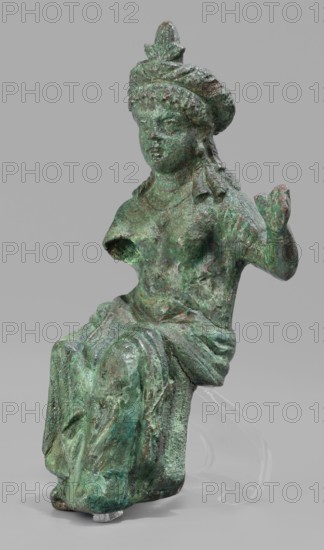 Figurine of the goddess Aphrodite-Isis, Egyptian deities, wigs, crowns, female figurines, Aphrodite mythology, Isis mythology, Egyptian art, 1st–2nd century, Roman period, Egypt ? Rome?, figurine, height 11.1 cm, width 5.3 cm, depth 5 cm, A green and brown weathered metal figure seated with draped clothing, showing a few areas of bright green patina and darker brown corrosion; the figure wears a rounded headdress with a pointed central element and has textured hair or a wreath around the head; the right arm is raised with a partially closed hand while the left arm is broken off near the shoulder; the torso shows worn surfaces and the legs are covered by drapery gathered over a block-like seat; the object rests against a neutral pale gray background and is supported by a transparent stand visible behind the lower portion.