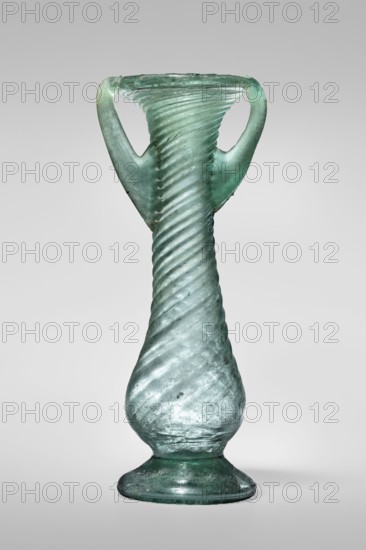 Unguentarium, unknown Eastern workshop. Glass vessel, transparent light green, Barag type VII: 1, 3. Unguentarium with pear-shaped baggy belly and neck decorated with diagonal ribs over the entire surface; foot with a gap; two vertical vices attached at two-thirds of the vessel's height and extending to the edge of the funnel-shaped rim. Free-blown. Outer rim diameter 4.50 cm; height 12.50 cm; outer bottom diameter 4.30 cm; weight 0.072 kg. 3rd–4th century, Syria., A translucent glass vessel with a twisted, spiral texture running vertically along its body, tapering toward a narrower neck and flaring slightly at the rim, featuring a pair of curved handles that rise from the shoulder to the rim; the glass shows a greenish-blue hue with areas of paler and darker shading, a few cloudy inclusions, and some surface irregularities, all set against a plain light gray background.