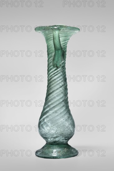 Unguentarium, unknown Eastern workshop. Glass vessel, transparent light green, Barag type VII: 1, 3, 3rd–4th century, Syria. Pear-shaped baggy belly and neck decorated with diagonal ribs over entire surface; foot with a gap. Two vertical vices attached at two-thirds of the vessel's height and extending to the edge of the funnel-shaped rim; free-blown. Outer rim diameter 4.50 cm; height 12.50 cm; outer bottom diameter 4.30 cm; weight 0.072 kg., A tall, narrow glass vessel with a flared rim and a rounded base on a short foot, made of translucent greenish-blue glass with a spiral ribbed texture running up the body; the glass surface shows slight iridescence and areas of cloudiness, with a thicker handle-like strand attached near the top that follows the curve to the rim, and the overall background is a smooth light gray.