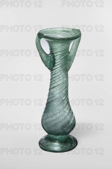 Unguentarium, unknown Eastern workshop. Glass vessel, transparent light green, Barag type VII: 1, 3. 3rd–4th century, Syria. Pear-shaped baggy belly and neck decorated with diagonal ribs over the entire surface; foot with a gap. Two vertical vices attached at 2/3 of the vessel's height and extended to the edge of the funnel-shaped rim; free-blown. Outer diameter of rim: 4.50 cm. Height: 12.50 cm. Outer diameter of bottom: 4.30 cm. Weight: 0.072 kg., A translucent green glass vessel with a spiraled, elongated body that widens toward a rounded base and narrows at the neck, topped by a flared rim; the surface shows helical ridges and areas of weathered texture. A pair of curved handles rise from the shoulder to the rim, each joining the body with irregular, slightly rough attachments. The glass displays variations of green and pale blue-green tones, with some darker green accents and subtle cloudy or frosted patches. The object stands against a pale neutral background.
