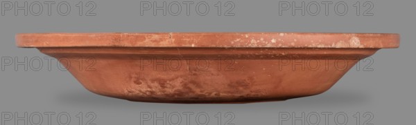 Bowl with palm leaf decoration. Fine-grained clay, pink-brown 2.5YR 5/6; glaze covers the entire surface, brown-red 2.5YR 4/8–5/8 with a slight sheen. Entire vessel slightly cracked; inside shows slight reflections of the glaze. In the center of the bottom is stamped decoration: four palmettes and four triple rings in a radial arrangement, surrounded by a roulette band between two concentric fluted grooves forming a medallion. Vessel made using a thin-walled technique, smooth at the fracture, classified as Group A Late Roman African pottery Late Roman Red Slip Ware A. Stamps: palmette Hayes type 10; triple rings Hayes type 26; stamped decoration in Palm Style B. Dated 360–450. Place of origin: Carthage Tunisia, Africa Proconsularis. Vessel, weight 0.824 kg; bowl height 5.9 cm, diameter 32.8 cm., A shallow, wide terracotta dish seen from the side with a rounded rim and a gently sloping body, showing surface wear and irregular patches of lighter discoloration; the dish is mainly reddish-...