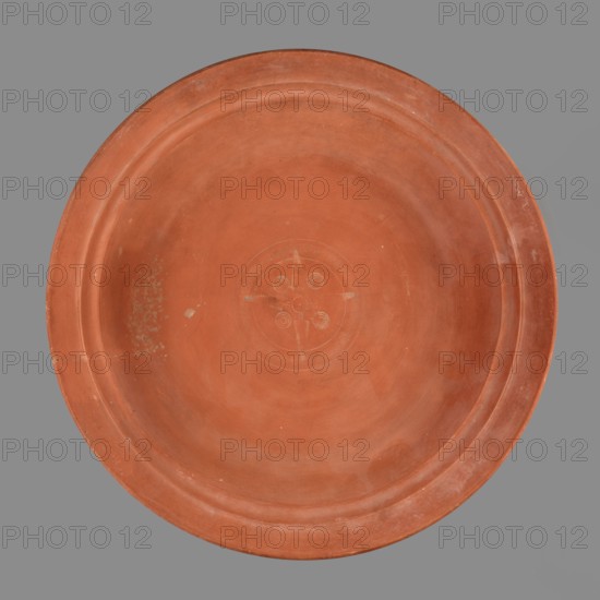 Bowl with palm leaf decoration. Roman pottery, Late Roman Red Slip Ware A Late Roman African pottery, ca. 360–450. Fine-grained clay, pink-brown 2.5YR 5/6; brown-red glaze 2.5YR 4/8–5/8 covering entire surface with slight sheen and slight cracking; inside shows slight glaze reflections. Thin-walled technique, smooth fracture Group A. In center of the bottom: stamped decoration forming a medallion — four palmettes and four triple rings in radial arrangement, surrounded by a roulette band between two concentric fluted grooves. Stamps: palmette Hayes type 10; triple rings Hayes type 26; stamped decoration classified as Palm Style B. Provenance: Carthage Tunisia, Africa Proconsularis. Vessel bowl weight 0.824 kg; height 5.9 cm; diameter 32.8 cm., A large round shallow dish viewed from above with a smooth terracotta surface in shades of reddish-brown and orange, showing a slightly raised rim and concentric circular indentations; the central area has a faint carved circular motif with a cros...