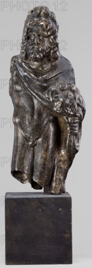 Figurine of Sylvanus in goat skin, with a pine wreath and a pine branch in his hand, figurine from a mold, Silvanus myth., 2nd century, Gaul France, figurine, height 23 cm, width 10 cm, depth 7.3 cm, weight 1.474 kg, A small sculpted figure of a robed person mounted on a rectangular block, shown from head to mid-legs; the figure has wavy hair and a beard, a draped garment across the chest, and holds a rounded bunch-like form in one hand; the surface of the sculpture is dark brown to black with lighter brown and greyish highlights and a slightly glossy, worn patina, while the supporting block is dark brown with subtle lighter brown spots; the background is a uniform light grey.