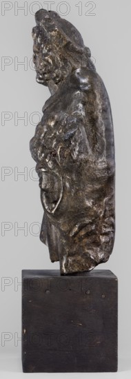 Figurine of Sylvanus in goat skin, with a pine wreath and a pine branch in his hand;, figurine from a mold; Silvanus myth., 2nd century, Gaul France; height 23 cm, width 10 cm, depth 7.3 cm, weight 1.474 kg, A three-dimensional dark metallic sculpture of a human bust shown in profile, mounted on a rectangular dark wooden block, with the sculpture displaying textured, uneven surfaces and a patina that ranges from deep brown to black with highlights of reflective bronze and muted gold, the figure's hair and facial features are roughly modeled with visible tool marks and irregular contours, the clothing or drapery suggested by folds and bulges continues down to a rough lower edge, the wooden base is matte dark brown with small lighter scuffs and subtle grain variations, and the background is a neutral pale gray.