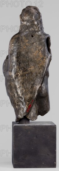 Figurine of Sylvanus in goat skin, with a pine wreath and a pine branch in his hand, figurines from a mold, Silvanus myth., 2nd century, Gaul France, figurine, height 23 cm, width 10 cm, depth 7.3 cm, weight 1.474 kg, A weathered sculptural form mounted on a rectangular block base, shown from the back. The sculpture has an irregular, organic surface with a draped, cloak-like outline and a rounded top suggesting a head. The surface shows varied textures with rough, pitted areas and smoother worn patches. Visible colors include dark brown, black, and patches of muted golden-brown and gray on the sculpture, while the base is a matte dark brown to black. There are a few small holes and indentations on the sculpture and an opening at one side, and the base shows subtle wear marks and small circular fastenings.