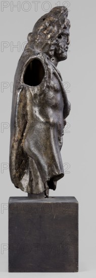 Figurine of Sylvanus in goat skin, with a pine wreath and a pine branch in his hand; unknown creator; figurine from a mold; Silvanus myth.; 2nd century; Gaul France; figurine; height 23 cm, width 10 cm, depth 7.3 cm, weight 1.474 kg, A dark bronze-colored sculpted figure shown in profile standing on a simple rectangular dark wooden base, with a weathered metallic surface that reflects hints of brown and muted gold, a textured cloak draped over the shoulders revealing a muscular torso and an exposed hollow shoulder area, a detailed head with curly hair and a bearded face turned slightly forward, irregular surface patina and small pits visible across the sculpture, and a neutral light gray background.