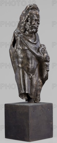 Figurine of Sylvanus in goat skin, with a pine wreath and a pine branch in his hand, figurines from a mold, Silvanus myth., 2nd century, Gaul France, figurine; height 23 cm, width 10 cm, depth 7.3 cm, weight 1.474 kg, A small sculpted figure mounted on a rectangular base, shown full-length from front; the figure appears to be a standing male with long hair and a beard, wearing a draped cloak that covers one shoulder and falls to mid-thigh, with a rolled or folded section near the neckline; one arm is bent and holds a rounded object against the chest while the other arm is partly obscured by the cloak and has an open hollow at the shoulder; the surface of the figure has a dark metallic patina with areas of lighter worn highlights in brownish and gray tones and patches of darker almost black coloration; the base is a plain block with a dark brown to black finish and visible small marks and tiny holes on its sides; the background is a uniform light gray.