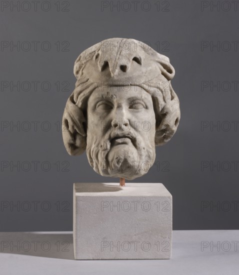 Head of Dionysus, unknown Italian workshop, 2nd half of the 1st century, Italy, sculpture, A weathered stone sculpted head of a bearded man mounted on a pale rectangular plinth, shown against a smooth gray background; the sculpture has rough, light gray and off-white tones with subtle darker gray shadows in the crevices, the beard and hair are carved with flowing, textured forms and the head is topped by a draped, rounded headdress with curved folds, creating deeper gray areas; the plinth appears creamy off-white with a slightly grainy surface and casts a soft shadow on the flat pale surface beneath.