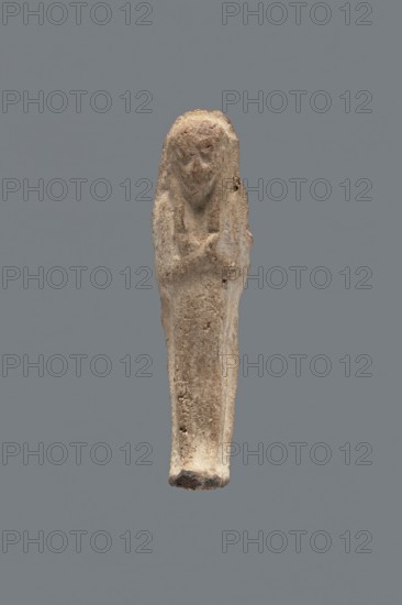 Shabti, mummy-shaped figurine, funerary gift, hoes; made carelessly, impressed in a mold. Hands folded on the chest, right over left; both hands hold hoes. Wears a three-part smooth wig. Facial details poorly legible. No inscription. Back unfinished. c. 1069–c. 656 BC, Third Intermediate Period, Egypt. Height 7.5 cm., A small elongated carved figure against a solid gray background, rendered in rough textured beige and light brown material with traces of darker brown in crevices; the figure has a rounded top suggesting a head with an indistinct facial area and a narrow tapering body, a subtle suggestion of crossed arms at chest level, and an uneven, slightly darker base; surface shows pitting and irregularities and a few small holes, with overall colors including beige, light brown, darker brown, and gray.