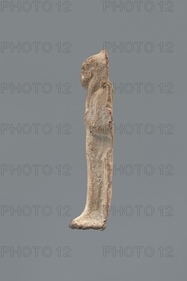 Shabti, Mummy-shaped figurine shabti, funerary gift; made carelessly, impressed in a mold. Hands folded on the chest, right over left; both hands hold hoes. Wears a three-part smooth wig. Facial details poorly legible. No inscription. Back unfinished. c. 1069–c. 656 BC, Third Intermediate Period, Egypt. Height 7.5 cm., A vertical, elongated object resembling a narrow carved figure displayed against a solid gray background, with a rough, weathered surface in shades of beige, tan, light brown, and hints of reddish-brown, showing irregular contours and a rounded top, a slightly curved midsection, and a broader, textured base that tapers into a small flat foot-like end; the surface has visible pits, small raised areas, and subtle color variations from pale cream to darker brown, with a faintly darker edge along one side.