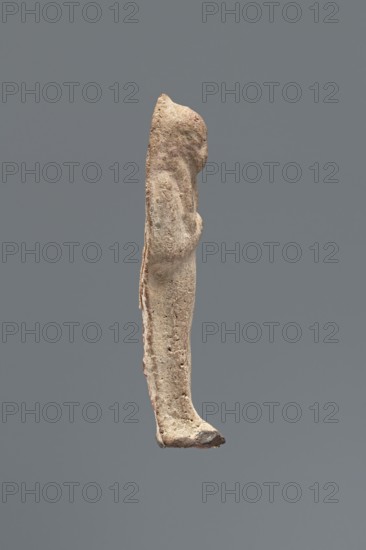 Shabti, Mummy-shaped funerary figurine, made carelessly and impressed in a mold. Hands folded on the chest, right over left; both hands hold hoes. Wears a three-part smooth wig. Facial details poorly legible. No inscription; back unfinished. Third Intermediate Period, Egypt, c. 1069–c. 656 BC. Shabti, height 7.5 cm., A vertically oriented, weathered beige figurine shown in profile against a solid gray background, with a rounded head, an elongated torso, and a slightly protruding arm positioned near the chest; the surface appears rough and pitted with areas of darker brown and faint reddish pigment along the back and lower edge, and a small darker spot near the base of the foot.