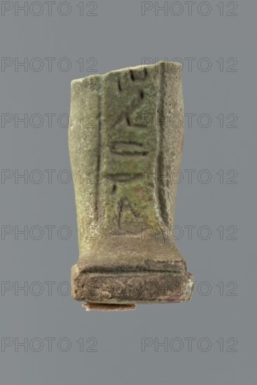 Shabti legs, mummy-shaped funerary figurine fragment: only the legs preserved from the knees down. The feet rest on a base with a flat pillar at the back. Inscribed text, preserved in fragments, placed in a column on the front of the legs and on the pillar. Late Period, Egypt, 664–332 BC. Shabti, height 4.2 cm., A small weathered object with a rough, uneven surface is shown against a plain gray background; the object is predominantly green with areas of brown and darker green patina, and it has a rectangular base that appears more brown and dusty with some lighter worn edges; the upper portion rises irregularly and shows vertical grooves and shallow impressed marks, with mottled patches of darker and lighter green and hints of tan; the edges are chipped and rough, and the overall appearance suggests aged metal or mineral with textured, corroded surfaces.