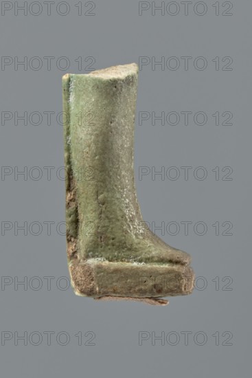 Shabti legs, Mummy-shaped funerary figurine fragment: legs preserved from knees down. Feet rest on a base with a flat pillar at the back. Inscribed text preserved in fragments in a column on the front of the legs and on the pillar. Late Period, Egypt, 664–332 BC. Shabti; height 4.2 cm., A small weathered object shaped like a short boot or shoe, shown in profile against a gray background; the object has a rough, pitted surface with a predominantly green tone mixed with areas of beige and brown, with some lighter whitish patches and darker brown spots, and appears to rest on a thicker rectangular base with a bit of frayed fibrous material visible beneath.