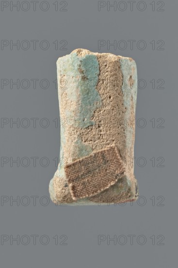 Shabti legs, mummy-shaped funerary figurine fragment: fragment of the figurine's legs preserved from the knees down; feet supported on a base with a flat pillar at the back. Shabti funerary gift, hoe, 664–332 BC Late Period?, Egypt. Height 3.9 cm., A roughly cylindrical object with a worn, rough surface, showing patches of light turquoise and sandy beige; the top edge is irregular and crumbled, and a small rectangular piece of coarse brownish fabric or woven material is affixed near the lower middle; the background is a solid mid-gray, and the object shows texture variations with areas of smoother pale turquoise and grainy beige erosion.