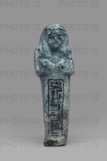 Ushabti Amenmose, Warmian-Masurian Voivodeship. Hieroglyphs; funerary figurine mummy-shaped shabti. Arms crossed on the chest, right over left. Holds hoes in both hands. Basket on the back. Smooth tripartite wig with headband; ears uncovered. Large eyes; mouth and nose not distinct. Tools, facial details, and headband painted with dark paint. Painted inscription in a framed column: Osiris the prophet of Amun Amenmose, justified by the voice. c. 1069–c. 656 BC, Third Intermediate Period, Egypt. Height 16.4 cm, width 5.3 cm., A small standing figure with a rounded head and simplified facial features, arms crossed over the chest and hands touching the shoulders, rendered in a weathered blue-green material with darker blue and black markings and areas of lighter turquoise and pale green patina; the surface shows rough texture and small patches of beige and gray where the material is worn, and the background is a smooth neutral gray.