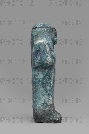 Ushabti Amenmose, Warmian-Masurian Voivodeship. Hieroglyphs; funerary figurine; wig; grave goods; mummy-shaped figurine. Arms crossed on the chest, right folded over left. Hoes in both hands. Basket on the back. Head with smooth tripartite wig and headband; ears uncovered. Large eyes; mouth and nose not distinct. Tools, facial details, and headband painted with dark paint. Painted inscription placed in a column of text enclosed in a frame: Osiris the prophet of Amun Amenmose, justified by the voice. c. 1069–c. 656 BC, Third Intermediate Period, Egypt. Shabti; height 16.4 cm, width 5.3 cm., The image shows a small vertical sculpture or figurine with a rough, pitted surface in a mix of blue, turquoise, green, and darker teal tones, with areas of lighter off-white and gray discoloration; the form stands on a flat base and has a simplified head and body profile with a rounded top and a subtle protrusion suggesting an arm or shoulder on one side; the background and surface beneath the objec...