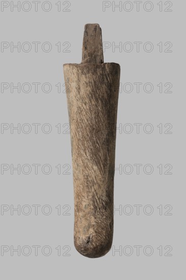 Beard from a sarcophagus mask, slightly curled; sarcophagus beard fragment, ca. 1550–332 BC New Kingdom–Late Period, Egypt; sarcophagus mask; height 16.2 cm., A single elongated wooden object with a rounded bottom and a squared top projection, showing a rough, weathered surface with visible grain and tool marks; the wood displays a range of brown tones from light tan to darker brown, with some grayish and muted greenish patches, against a uniform light gray background.