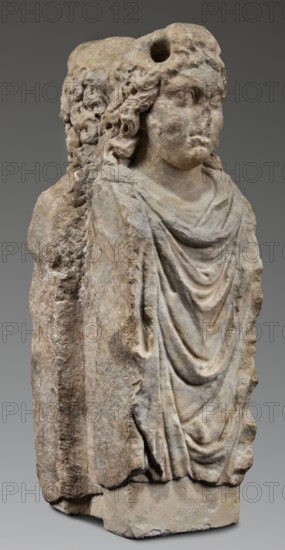 Double herm with heads of Apollo, unknown Roman workshop; herm; Apollo myth.; Roman sculpture, Antonine period 138–192 CE, Roman Empire 27 BC–476; sculpture; height 67 cm, width 27 cm, depth 24 cm, weight 85.5 kg, A carved stone figure with a weathered surface stands against a plain gray background; the sculpture shows a draped garment with flowing folds and a visible shoulder and neckline, and a head with curly hair and softened facial features, including a rounded brow, eyes, nose, and mouth. The stone displays a range of colors including pale beige, light gray, warm tan, and darker brown patches, with areas of lighter off-white on raised surfaces and some darker weathered spots in crevices. The back of the piece appears rougher and more irregular than the carved front, and the base is a block of the same stone showing chipping and uneven edges.
