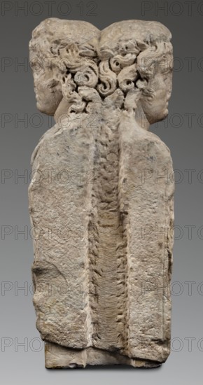 Double herm with heads of Apollo, unknown Roman workshop; sculpted Roman marble herm; subject: Apollo myth.; herm form; Roman sculpture, Antonine period 138–192 CE, Roman Empire 27 BC–476 CE; sculpture; height 67 cm, width 27 cm, depth 24 cm, weight 85.5 kg., A weathered stone sculpture showing the backs of two human heads joined at the back, each head turned slightly outward so profiles are visible on either side; the heads have carved curly hair with swirling locks, and the upper portion of the stone shows textured, eroded surfaces and rounded contours. The central area between the heads features a vertical recessed channel with a rough, ridged texture. The lower portion of the sculpture widens into a blocky base with uneven, chipped edges and pitted surfaces. Visible colors include pale beige and off-white in the stone, with areas of light gray and subtle darker gray discoloration, and small patches of yellowish-brown. The background behind the sculpture is a smooth gradient of medi...