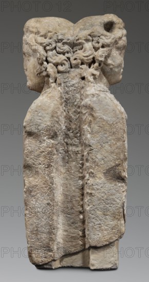 Double herm with heads of Apollo, unknown Roman workshop; herms; Apollo myth.; Roman sculpture, Antonine period 138–192; Roman Empire 27 BC–476; sculpture; height 67 cm, width 27 cm, depth 24 cm, weight 85.5 kg, The image shows a weathered stone sculpture of two human heads back-to-back atop a shared vertical block, with a rough textured surface and visible erosion. The heads have curly hair carved along the center where they meet, and both faces are shown in profile facing opposite directions. The stone displays a range of beige, tan, off-white, light brown, and gray tones, with darker gray shadows in crevices and lighter creamy areas on raised surfaces. The background is a smooth mid-gray.