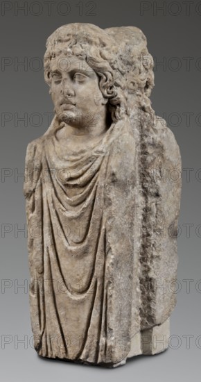 Double herm with heads of Apollo, unknown Roman workshop; herms; Apollo myth.; Roman sculpture, 138–192 Antonine period, Roman Empire 27 BC–476; sculpture; height 67 cm, width 27 cm, depth 24 cm, weight 85.5 kg., The image shows a weathered stone sculpture of a draped figure with a carved head and torso; the figure has wavy hair and a calm facial expression with closed or empty eyes, and the drapery falls in layered folds over the chest and down the front. The sculpture surface is rough and worn with visible chips and erosion, and there is a block-like extension at the back suggesting the figure remains attached to a larger stone. The colors visible are shades of beige, tan, light brown, gray, and off-white, with darker gray areas where the stone is more recessed or stained.