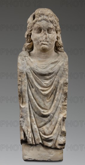 Double herm with heads of Apollo, unknown Roman workshop; herms; Apollo myth.; Roman sculpture; Antonine period 138–192; Roman Empire 27 BC–476; sculpture; height 67 cm, width 27 cm, depth 24 cm, weight 85.5 kg, A weathered stone sculpture of a human figure seen from the front, carved as a bust and torso on a rectangular base, with long wavy hair falling to the shoulders and a rounded face with softened features; the figure wears a draped garment displaying a group of layered folds cascading down the chest and torso; the surface shows rough, pitted texture and areas of erosion. The visible colors are various shades of beige, light brown, gray, and off-white, with darker gray shadows in the drapery folds and subtle brownish discolorations across the stone.