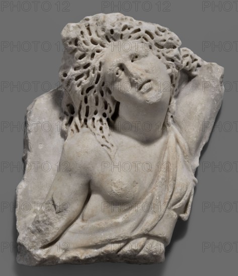 Fragment of a sarcophagus with a depiction of the Amazonomachy, unknown Roman workshop, 2nd quarter of the 3rd century, Roman Empire 27 BC–476, sarcophagus, height 35 cm, width 31 cm, depth 12 cm, weight 13 kg, A carved stone fragment showing the torso and head of a reclining female figure with flowing, perforated hair, textured surface, and an exposed shoulder and breast partially covered by draped fabric; the stone appears pale beige to off-white with subtle areas of darker discoloration and weathering, and the background is a solid medium gray.