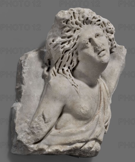 Fragment of a sarcophagus with a depiction of the Amazonomachy, unknown Roman workshop, 2nd quarter of the 3rd century, Roman Empire 27 BC–476, sarcophagus; height 35 cm, width 31 cm, depth 12 cm, weight 13 kg, A weathered stone sculpture fragment depicting an upper torso and head of a figure leaning back with a tilted face and open mouth; the figure has textured, carved hair with many elongated holes and strands, a visible shoulder and chest with draped cloth across the body, and the stone shows worn, rough surfaces and chips; the colors visible are shades of off-white, light gray, pale beige, and touches of darker gray.