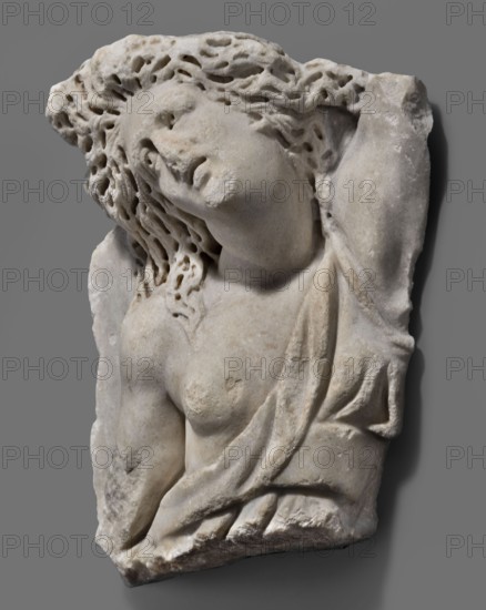 Fragment of a sarcophagus with a depiction of the Amazonomachy, unknown Roman workshop, 2nd quarter of the 3rd century, Roman Empire 27 BC–476, sarcophagus; height 35 cm, width 31 cm, depth 12 cm, weight 13 kg, A carved pale stone relief of a reclining female figure with wavy, textured hair, a tilted head and closed eyes, showing a bare torso partially covered by draped cloth; the stone surface shows subtle variations in off-white, beige and light gray tones, with darker gray shadowing and some brownish discoloration in places, set against a neutral medium gray background.