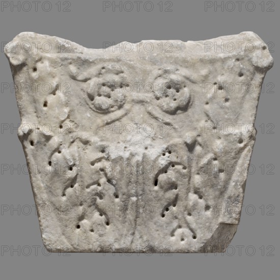 Corinthising pilaster capital, Corinthising style; architectural element / capital; decorative motifs: acanthus leaves, rosettes; function: element of architectural decoration; material/technique: Roman architecture; date: 1st–2nd century turn of the 1st and 2nd centuries; place of origin: Roman Empire 27 BC–476; dimensions: height 39 cm, width 46.5 cm, depth 11 cm; weight 36 kg., A roughly rectangular carved stone block with an irregular top edge and worn surfaces, showing a pale off-white to light gray color with areas of slightly darker gray and subtle beige tones; the front face features shallow relief motifs including a pair of circular, rosette-like shapes near the top connected by a low-relief band, and a vertical, central fluted or ribbed element below them flanked by a few curved, leaf-like forms; the surface has numerous small rounded pits and holes, weathered edges, and a few chipped corners, all set against a neutral gray background.