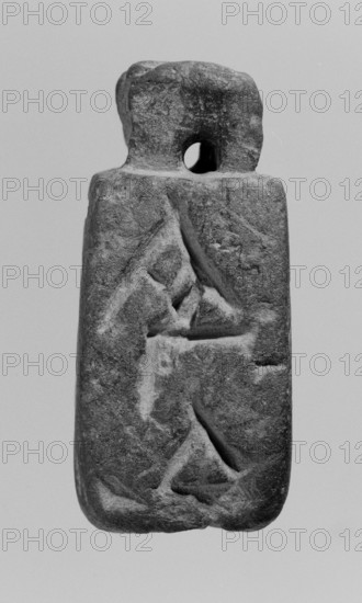 Christian amulet with Greek inscription. Limestone amulet in the shape of an elongated hexagon. There is text in Greek on the sides and on the bottom. On the upper edge, there is a protrusion with a hole drilled through it, most likely used to attach a strap or chain. The letters on the five mentioned edges form a coherent inscription in Greek with magical meaning. 2nd–3rd century, Miletus place of finding. Amulet. Height 5.2 cm, width 2.3 cm., A vertically oriented stone object with a rough texture and a rectangular lower portion topped by an irregular upper section with a rounded hole near the top; the surface shows carved shallow grooves forming angular shapes and incised lines; the stone displays a range of grayscale tones from light gray to dark gray with midtone variations; the background is a uniform pale gray.