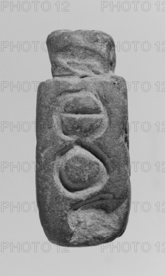 Christian amulet with Greek inscription. Limestone amulet in the shape of an elongated hexagon. There is text in Greek on the sides and on the bottom. On the upper edge, there is a protrusion with a hole drilled through it, most likely used to attach a strap or chain. The letters on the five mentioned edges form a coherent inscription in Greek with magical meaning. 2nd–3rd century, Miletus findspot, amulet, height 5.2 cm, width 2.3 cm., The image shows a vertically oriented carved stone object with a rough texture and visible wear; its surface displays carved circular and rounded motif shapes arranged along its length, and the stone appears in shades of gray and darker gray against a pale gray background.